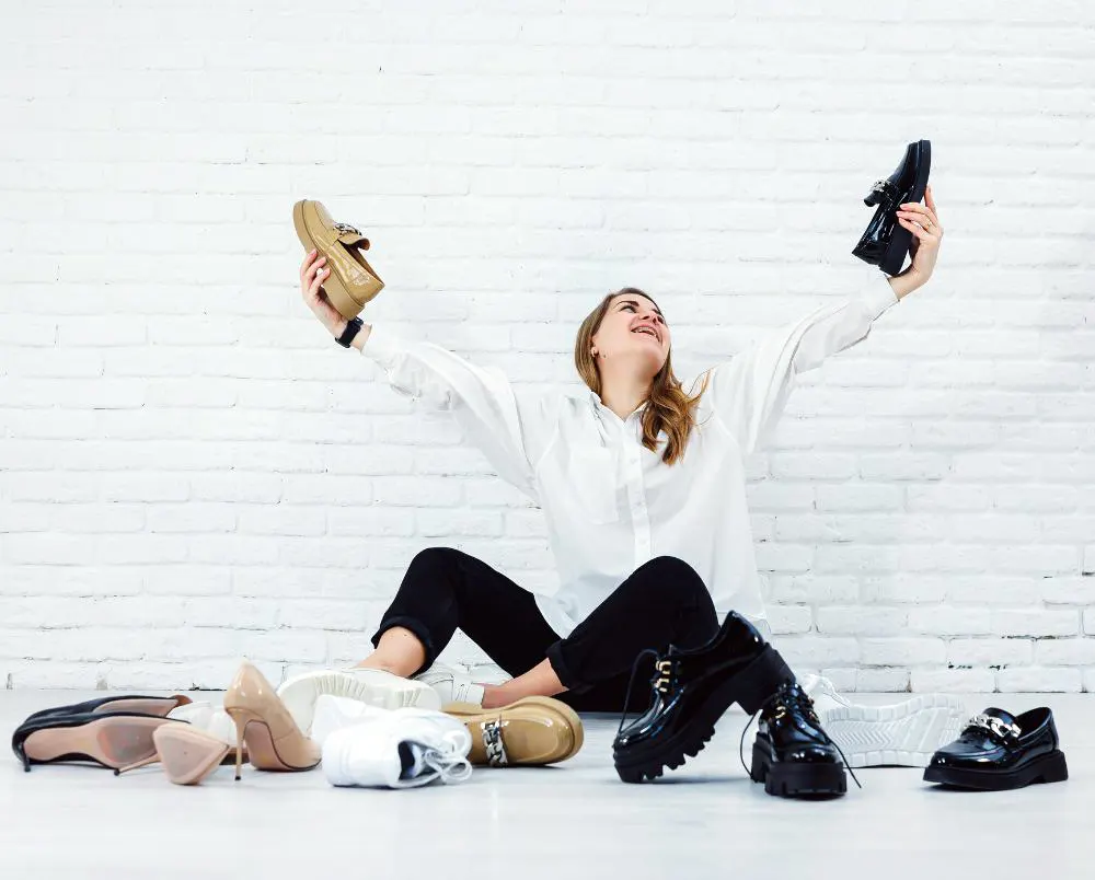 Woman with a smile in a fashionable outfit with a white shirt and black trousers sits on the floor among the shoes and chooses a new pair of shoes for herself